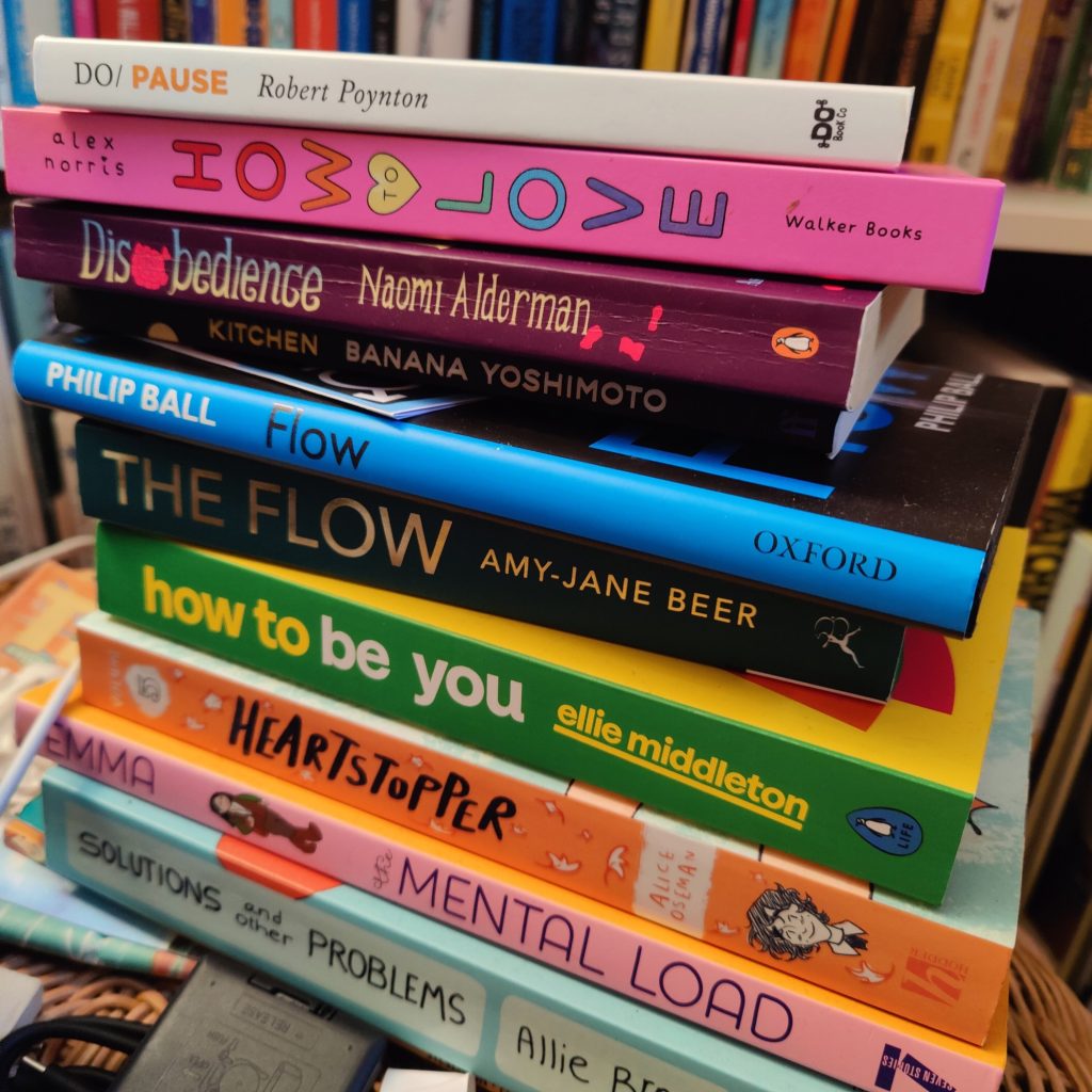 A slightly unstable stack of books. We can see their spines:
DO/PAUSE, by Robert Poynton
HOW to LOVE, by Alex Norris
Disobedience, by Naomi Alderman
Kitchen, by Banana Yoshimoto
Flow, by Philip Ball
The Flow, by Amy-Jane Beer
how to be you, by ellie middleton
Heartstopper (vol 5) by Alice Oseman
Mental Load, by Emma
Solutions and Other Problems, by Allie Brosh