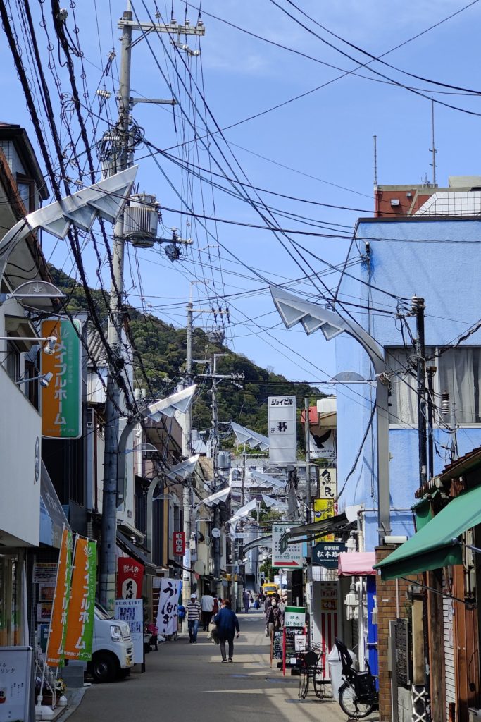 A quiet, yet visually busy Japanese street: many overhead wires and signs, a verdant hill in the background, and a few indistinct humans looking small by comparison.