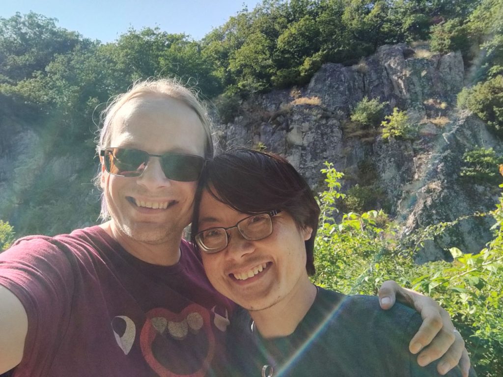 Fergus (a white person with long, blonde-ish hair, wearing sunglasses and a red t-shirt with one of AJ Simpson's Blob faces) and Sonny (who is half-Chinese, wearing glasses and a grey-green t-shirt). We are standing in front of a wooded cliff, looking happy.
